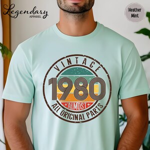 May include: A light green t-shirt with a vintage 1980 graphic. The graphic is a circular design with the word "VINTAGE" at the top, the number "1980" in the center, and the words "ALL ORIGINAL PARTS" at the bottom.