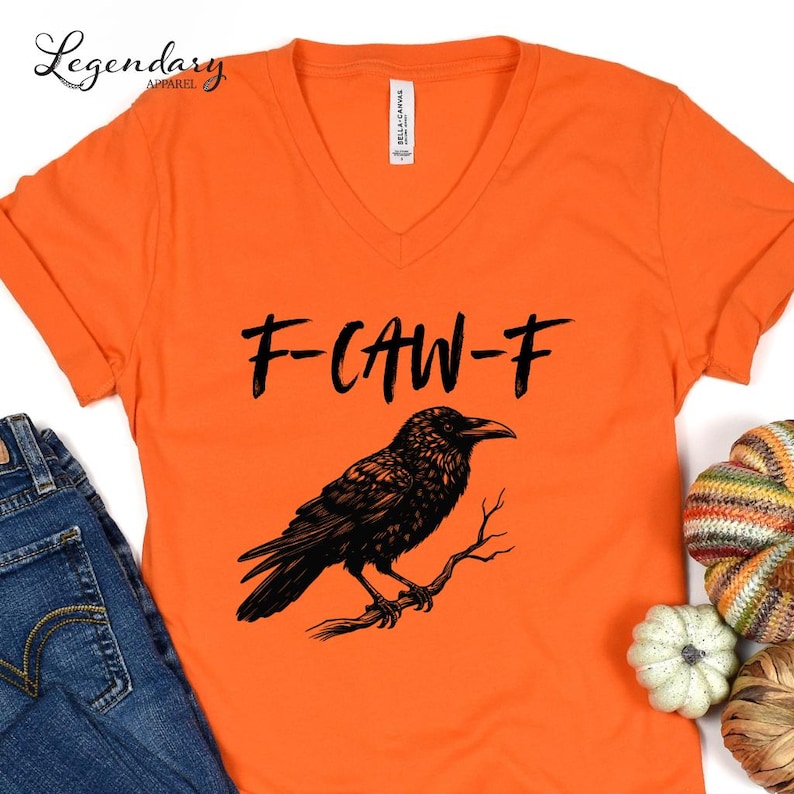 Pode incluir: Camiseta laranja com decote em V com o texto "F-CAW-F" e uma ilustra&ccedil;&atilde;o de um corvo preto empoleirado em um galho. A camiseta &eacute; feita de um material macio.