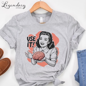 Pu&ograve; includere: Una maglietta grigia con un'illustrazione retr&ograve; di una donna che tiene in mano un cervello, con la scritta "USE IT!". Il design presenta uno stile vintage con uno sfondo color corallo.