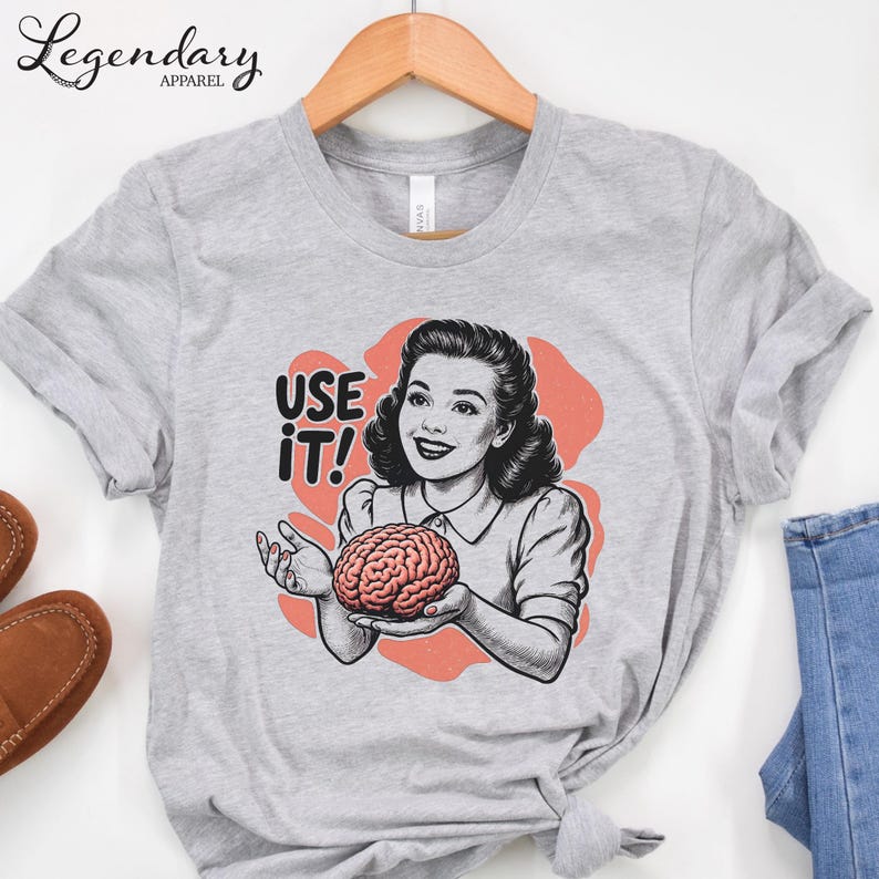 Pu&ograve; includere: Una maglietta grigia con un'illustrazione retr&ograve; di una donna che tiene in mano un cervello, con la scritta "USE IT!". Il design presenta uno stile vintage con uno sfondo color corallo.
