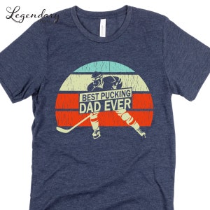 May include: A navy blue t-shirt with a vintage-style graphic of a hockey player holding a hockey stick and a sign that says "Best Pucking Dad Ever".