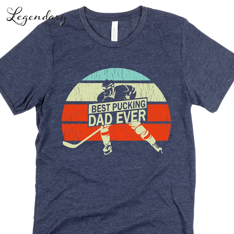 May include: A navy blue t-shirt with a vintage-style graphic of a hockey player holding a hockey stick and a sign that says "Best Pucking Dad Ever".