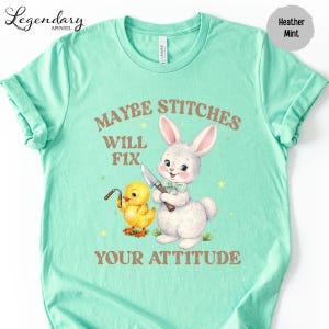 A turquoise t-shirt with a cartoon rabbit and a yellow duck holding a knife, accompanied by the text "MAYBE STITCHS WILL FIX YOUR ATTITUDE".
