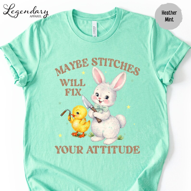 A turquoise t-shirt with a cartoon rabbit and a yellow duck holding a knife, accompanied by the text "MAYBE STITCHS WILL FIX YOUR ATTITUDE".
