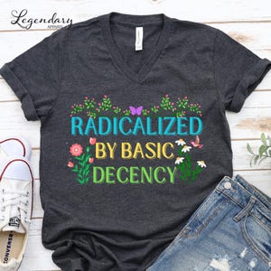May include: Dark gray V-neck t-shirt with the text "RADICALIZED BY BASIC DECENCY" in colorful letters. The design includes floral and butterfly accents. The shirt is paired with blue jeans and white sneakers.
