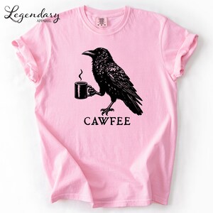 A pink t-shirt with a black illustration of a crow holding a coffee cup, accompanied by the text "CAWFEE".