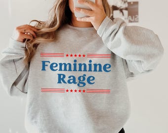 Könnte beinhalten: Ein grauer Pullover mit dem Text "Feminine Rage" in blauen Buchstaben mit roten Sternen und Streifen über und unter dem Text.