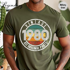 May include: Military green t-shirt with a vintage 1980 graphic. The graphic is a circular design with the text "VINTAGE 1980 ALL ORIGINAL PARTS" in white, with a retro color scheme of teal, yellow, orange, and red.