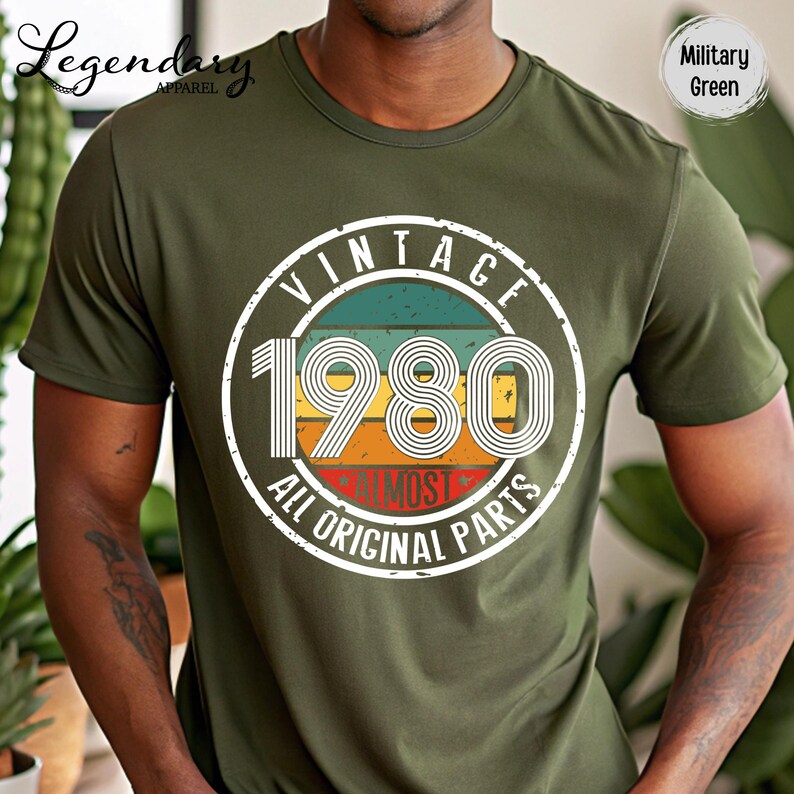 May include: Military green t-shirt with a vintage 1980 graphic. The graphic is a circular design with the text "VINTAGE 1980 ALL ORIGINAL PARTS" in white, with a retro color scheme of teal, yellow, orange, and red.