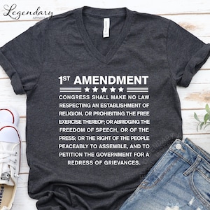 May include: Dark gray v-neck t-shirt with the text "1st Amendment" and the full text of the First Amendment of the United States Constitution in white. The shirt is paired with blue jeans and white sneakers.