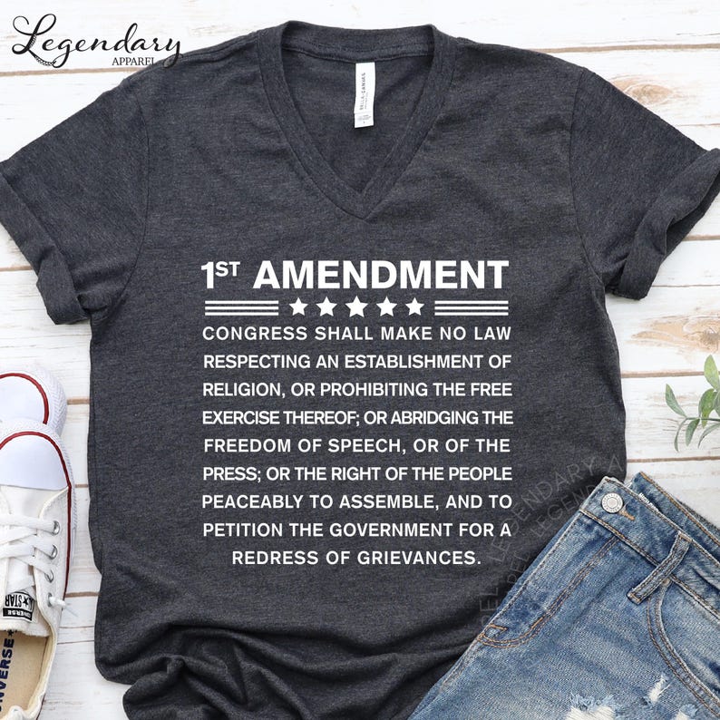 A gray t-shirt with white text printed on it, laid out on a wooden surface next to a pair of jeans.