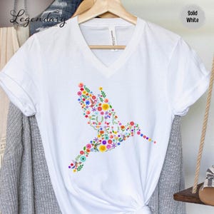 May include: White v-neck t-shirt with a colorful hummingbird design made of flowers. The hummingbird is facing right and is in flight.