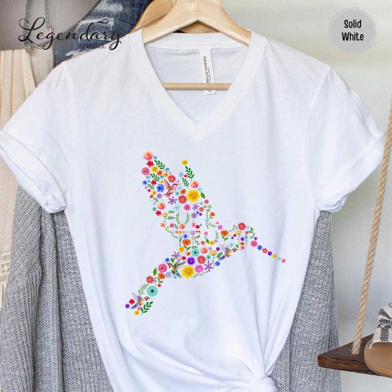 May include: White v-neck t-shirt with a colorful hummingbird design made of flowers. The hummingbird is facing right and is in flight.