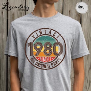 May include: Grey t-shirt with a vintage-style graphic. The graphic features the year "1980" in a retro font, surrounded by a circular design that reads "VINTAGE" and "ALL ORIGINAL PARTS". The shirt is from "Legendary Apparel".