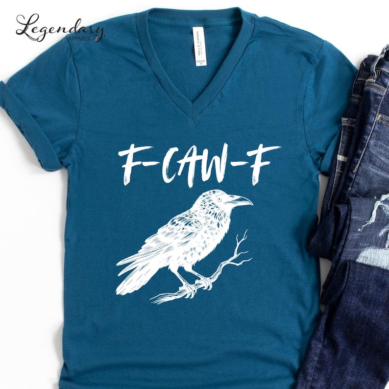 Pode incluir: Camiseta azul-petr&oacute;leo com decote em V com o texto "F-CAW-F" acima de uma ilustra&ccedil;&atilde;o branca de um corvo empoleirado em um galho. A camisa tem mangas enroladas e uma etiqueta "Bella + Canvas".
