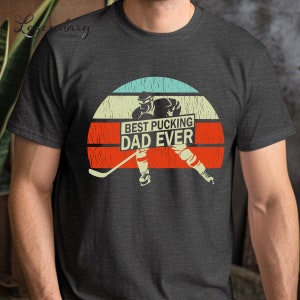 May include: A dark gray t-shirt with a vintage-style graphic of a hockey player skating with a hockey stick. The graphic is set against a red, orange, and blue sunset background. The text "BEST PUCKING DAD EVER" is displayed on a white banner.