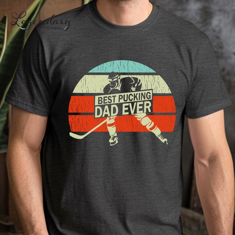 May include: A dark gray t-shirt with a vintage-style graphic of a hockey player skating with a hockey stick. The graphic is set against a red, orange, and blue sunset background. The text "BEST PUCKING DAD EVER" is displayed on a white banner.