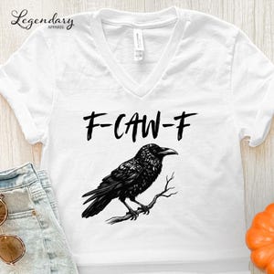 Pode incluir: Camiseta branca com decote em V com um gr&aacute;fico preto de um corvo empoleirado em um galho, e o texto "F-CAW-F" acima do p&aacute;ssaro. A camiseta &eacute; feita de um material macio.