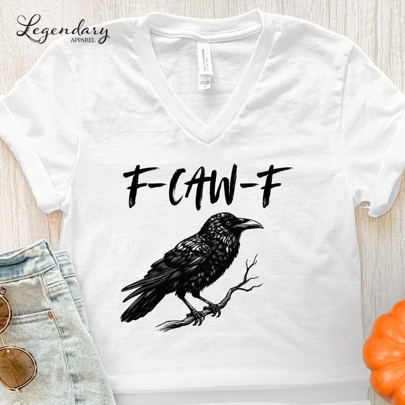 Pode incluir: Camiseta branca com decote em V com um gr&aacute;fico preto de um corvo empoleirado em um galho, e o texto "F-CAW-F" acima do p&aacute;ssaro. A camiseta &eacute; feita de um material macio.