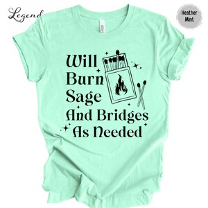 May include: A light green t-shirt with the text "Will Burn Sage And Bridges As Needed" in black. The text is accompanied by a graphic of a box of matches with a flame and two crossed matches.