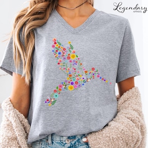 May include: A gray V-neck t-shirt with a floral hummingbird design. The hummingbird is made up of colorful flowers and leaves.