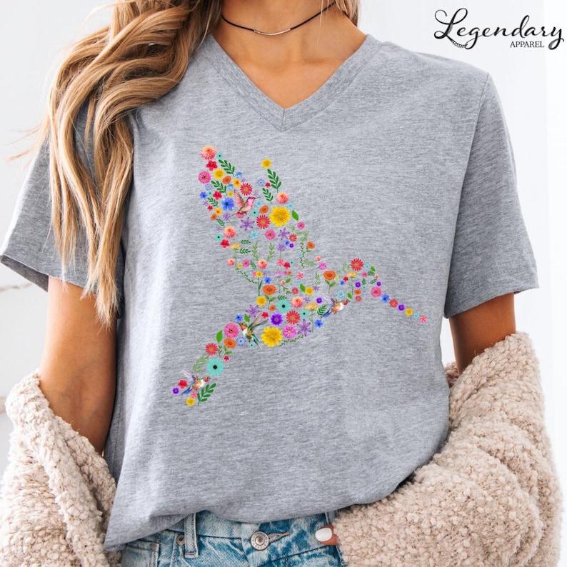 May include: A gray V-neck t-shirt with a floral hummingbird design. The hummingbird is made up of colorful flowers and leaves.