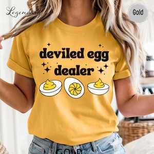 May include: A gold-colored t-shirt featuring the black text 'deviled egg dealer' in a rounded font. Below the text are three illustrations of halved deviled eggs with yellow yolks and white egg whites, accented by small black star symbols.