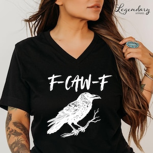 Pode incluir: T-shirt preta com decote em V com o texto branco "F-CAW-F" e uma ilustra&ccedil;&atilde;o detalhada de um corvo empoleirado num galho. A t-shirt &eacute; da Legendary Apparel.