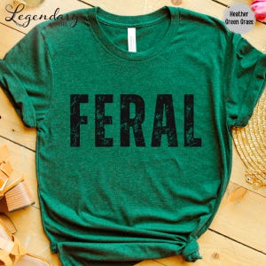 a green t-shirt with the word "FERAL" printed on it, laid out on a wooden surface.