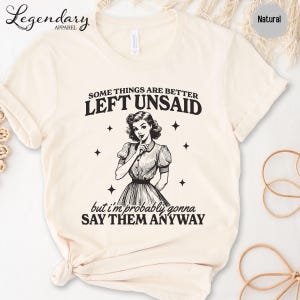 Może przedstawiać: Kremowa koszulka z napisem "SOME THINGS ARE BETTER LEFT UNSAID" i "but I'm probably gonna SAY THEM ANYWAY." Zawiera ilustrację w stylu vintage kobiety z palcem na ustach. Koszulka jest oznaczona jako "Natural".