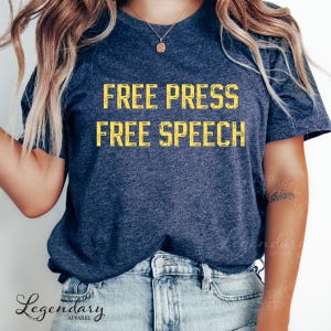 May include: Navy blue t-shirt with the text "FREE PRESS FREE SPEECH" in yellow block letters. The shirt is made of a soft material and is paired with blue jeans. The shirt is from Legendary Apparel.