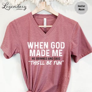 A maroon t-shirt with the text "WHEN GOD MADE ME" printed on it, along with the phrase "HE GRINNED AND SAID 'THIS'LL BE FUN'" printed below.