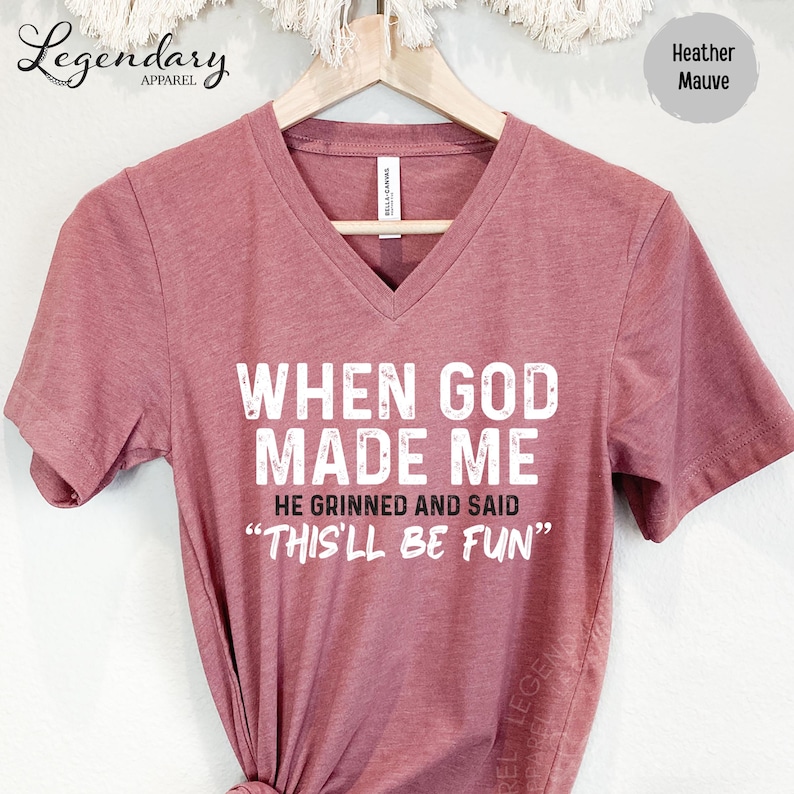 A maroon t-shirt with the text "WHEN GOD MADE ME" printed on it, along with the phrase "HE GRINNED AND SAID 'THIS'LL BE FUN'" printed below.