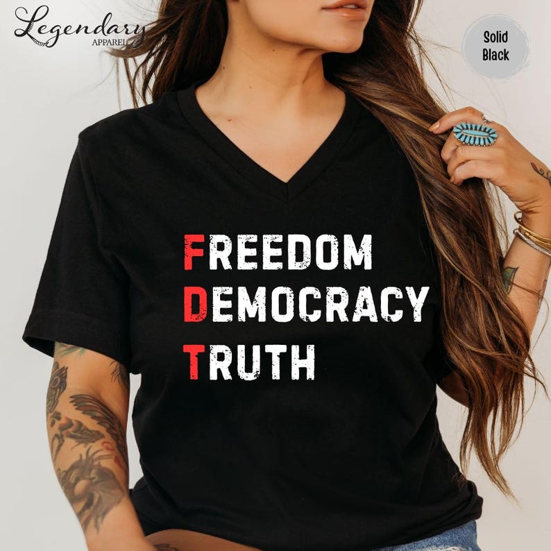 Op de afbeelding: Zwart V-hals T-shirt met de woorden "FREEDOM DEMOCRACY TRUTH" in wit en rood gedrukt. Het shirt is effen zwart van kleur en heeft het Legendary Apparel logo.