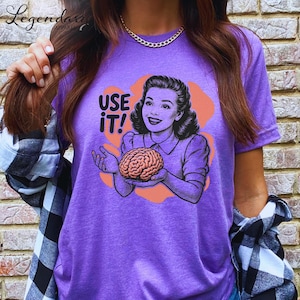 Pu&ograve; includere: Una maglietta viola con la scritta "USE IT!" e un'illustrazione vintage di una donna che tiene in mano un cervello. La grafica &egrave; in nero, arancione e marrone. La maglietta &egrave; a maniche corte.