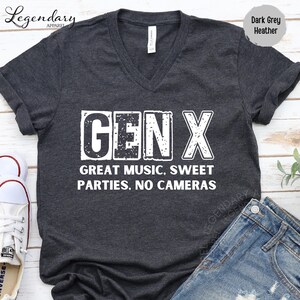 A gray t-shirt with the text "GENX" printed on it, along with the text "GREAT MUSIC. SWEET PARTIES. NO CAMERAS" printed below it. The t-shirt is laid out on a wooden surface, and there are some shoes and jeans visible in the background.