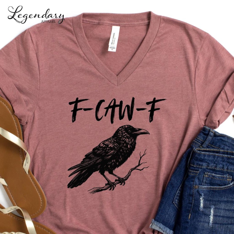 Pode incluir: Camiseta rosa empoeirada com decote em V com o texto "F-CAW-F" acima de uma ilustra&ccedil;&atilde;o de um corvo preto empoleirado em um galho. A camiseta &eacute; combinada com shorts jeans azuis.