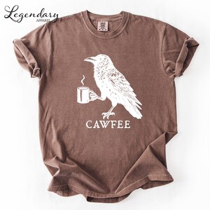 a brown t-shirt with a white graphic design depicting a bird holding a cup of coffee.