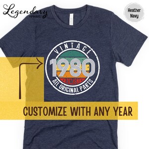 May include: Heather navy t-shirt with a circular graphic that reads "VINTAGE 1980 ALL ORIGINAL PARTS." The graphic has a retro design with green, orange, and red stripes. The text "CUSTOMIZE WITH ANY YEAR" is also visible.
