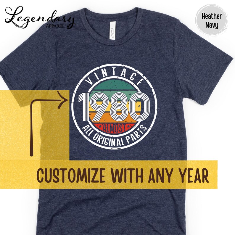 May include: Heather navy t-shirt with a circular graphic that reads "VINTAGE 1980 ALL ORIGINAL PARTS." The graphic has a retro design with green, orange, and red stripes. The text "CUSTOMIZE WITH ANY YEAR" is also visible.