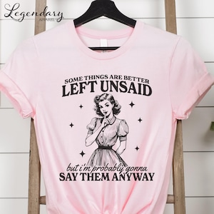 Może przedstawiać: R&oacute;żowa koszulka z napisem "SOME THINGS ARE BETTER LEFT UNSAID" i "but I'm probably gonna SAY THEM ANYWAY". Grafika przedstawia ilustrację w stylu vintage kobiety z palcem na ustach.