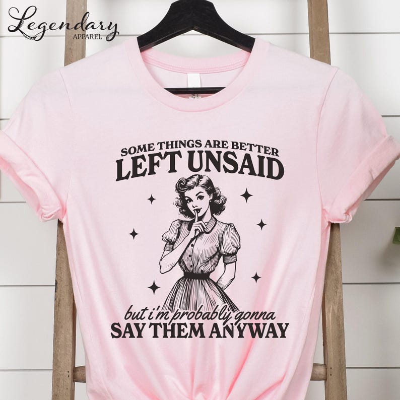 Może przedstawiać: R&oacute;żowa koszulka z napisem "SOME THINGS ARE BETTER LEFT UNSAID" i "but I'm probably gonna SAY THEM ANYWAY". Grafika przedstawia ilustrację w stylu vintage kobiety z palcem na ustach.