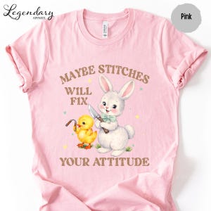 A pink t-shirt with a cartoon rabbit and a yellow duck holding a knife, accompanied by the text "MAYBE STITCHS WILL FIX YOUR ATTITUDE".