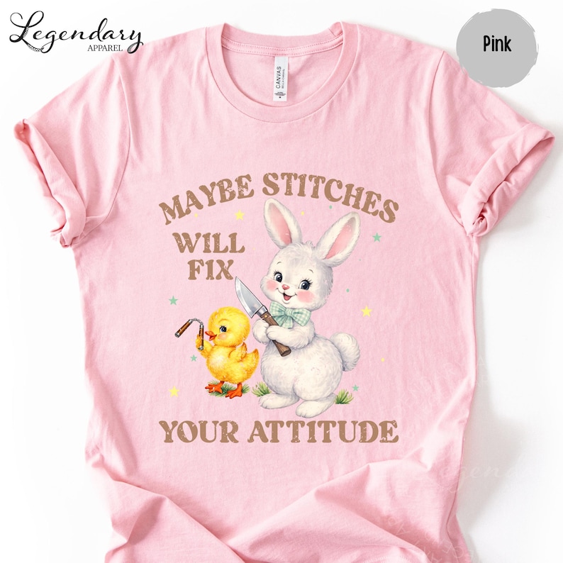 A pink t-shirt with a cartoon rabbit and a yellow duck holding a knife, accompanied by the text "MAYBE STITCHS WILL FIX YOUR ATTITUDE".