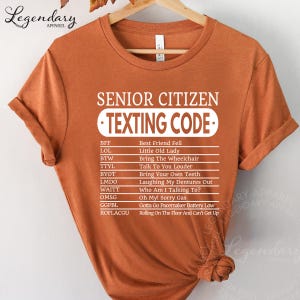 a t-shirt with a list of various texts, including "Best Friend Fell", "Little Old Lady", "Bring The Wheelchair", "Bring Your Own Teeth", "Laughing My Dentures Out", "OMM0", "Gotta Go Pancake Maker Battery Low", and "Rolling On The Floor And Can't Get Up".