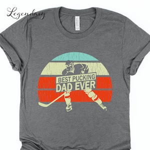 May include: A gray t-shirt with a vintage-style graphic of a hockey player holding a sign that says "Best Pucking Dad Ever".