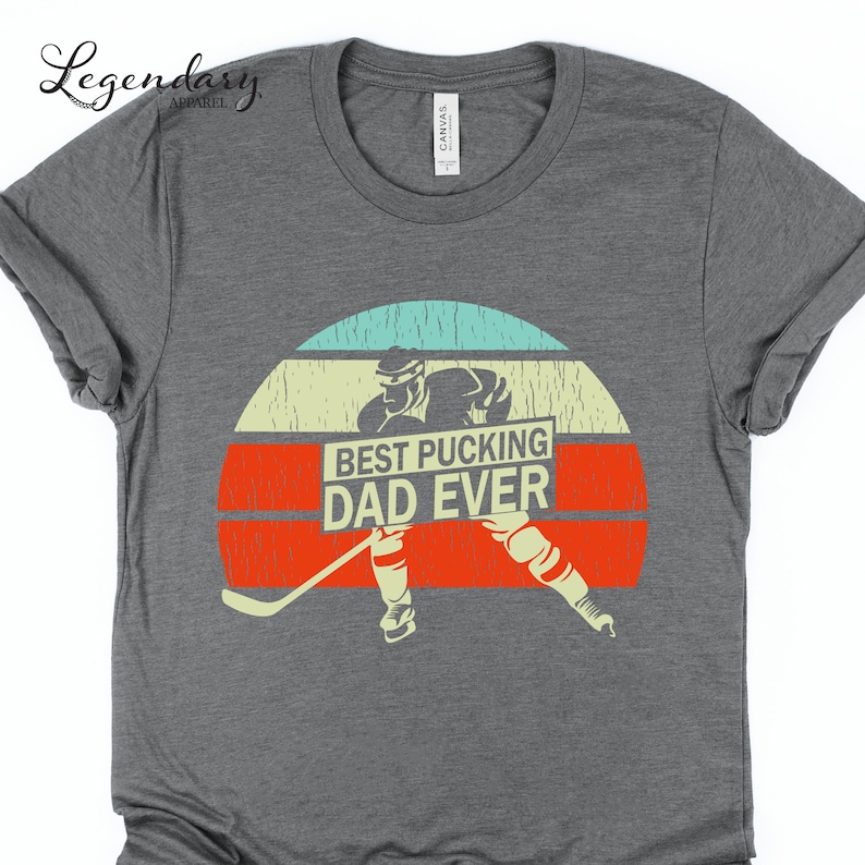 May include: A gray t-shirt with a vintage-style graphic of a hockey player holding a sign that says "Best Pucking Dad Ever".