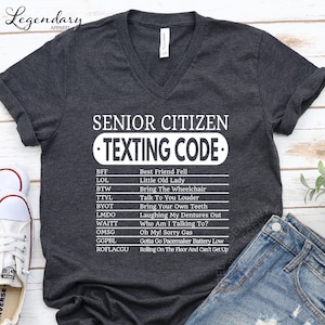 A gray t-shirt with a humorous text printed on it, along with a pair of jeans and a pair of sneakers, all laid out on a wooden surface.