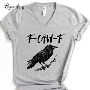 Pode incluir: Camiseta cinza claro com decote em V com uma ilustra&ccedil;&atilde;o de um corvo preto empoleirado em um galho. O texto "F-CAW-F" est&aacute; acima do corvo. A camiseta tem mangas curtas e uma etiqueta "Bella + Canvas". A marca "Legendary Apparel" est&aacute; no canto superior esquerdo.