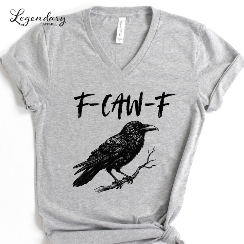 Pode incluir: Camiseta cinza claro com decote em V com uma ilustra&ccedil;&atilde;o de um corvo preto empoleirado em um galho. O texto "F-CAW-F" est&aacute; acima do corvo. A camiseta tem mangas curtas e uma etiqueta "Bella + Canvas". A marca "Legendary Apparel" est&aacute; no canto superior esquerdo.